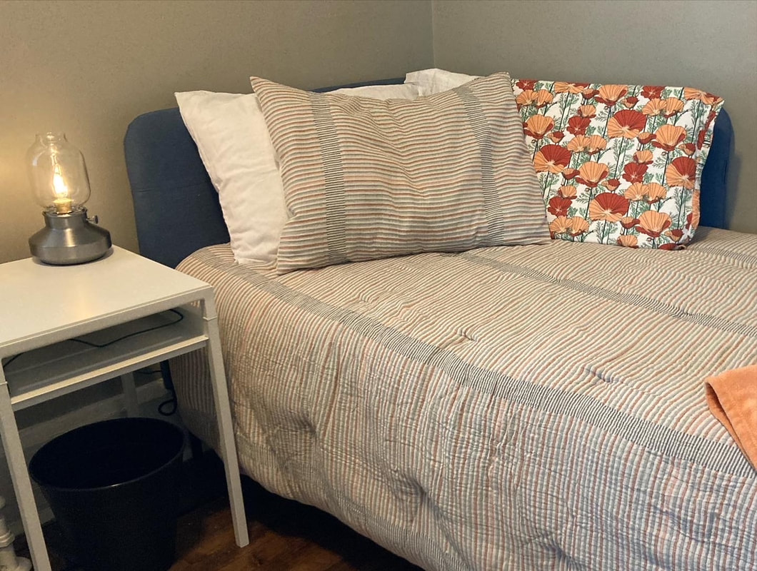 Image: A bed with a striped comforter, striped pillow and floral pillow. Next to the bed, there's a small white bedside table with a lamp.